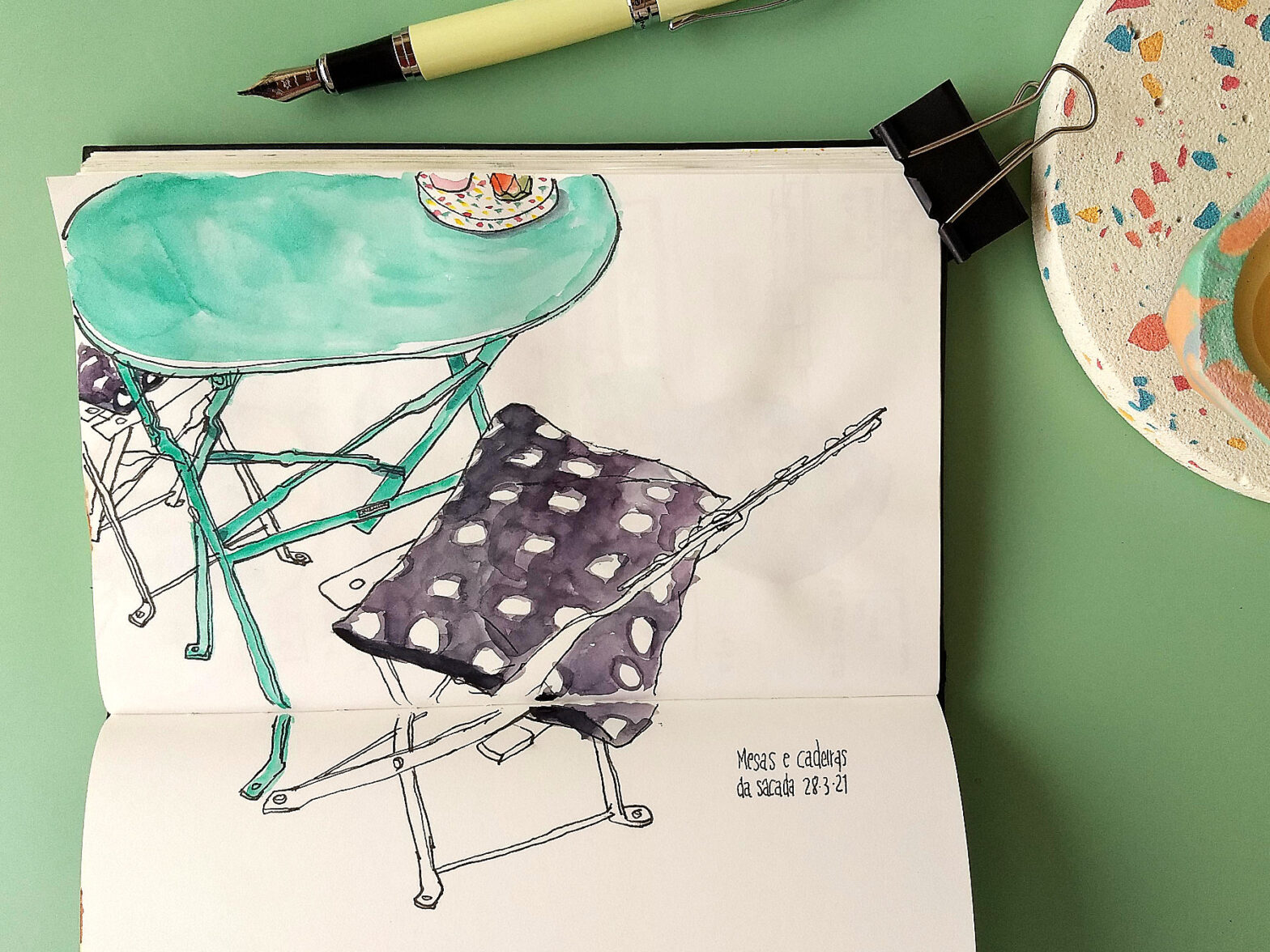 Caderno com desenho de uma mesa redonda com duas cadeiras, todas de metal. O caderno está rodeado por uma caneta e um suporte