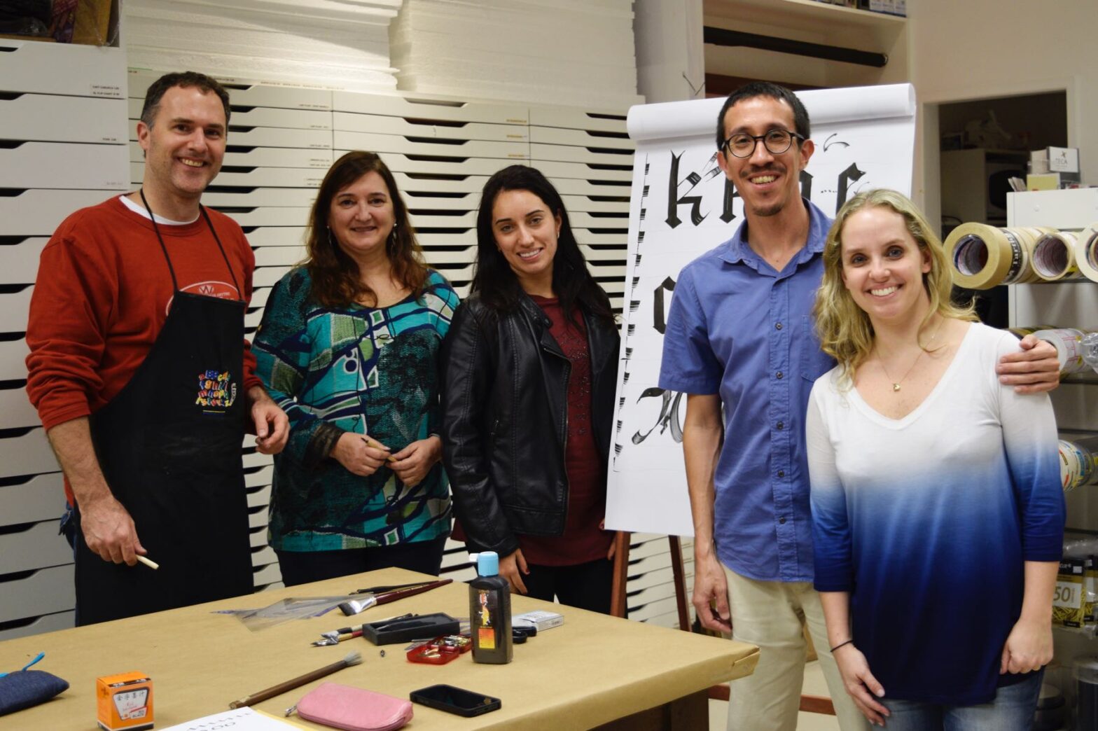 Foto com quatro participantes da oficina e o instrutor, junto à mesa e à frente de um flowchart com modelos de letras