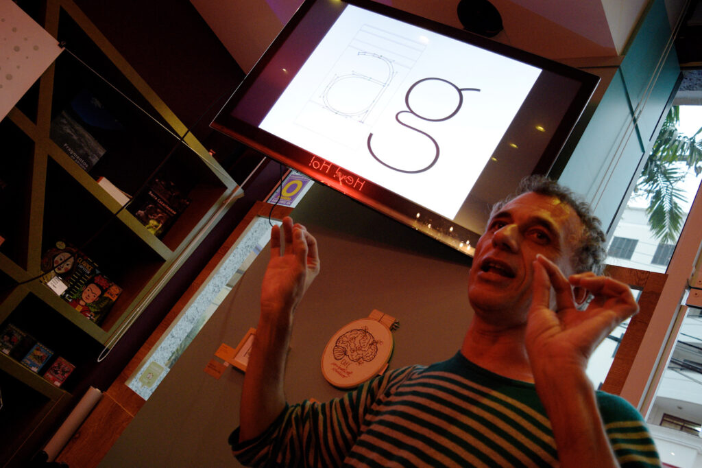 Jefferson Cortinove fala em frente a uma tela de TV mostrando detalhes da letra “g” no Coffee & Shop 18