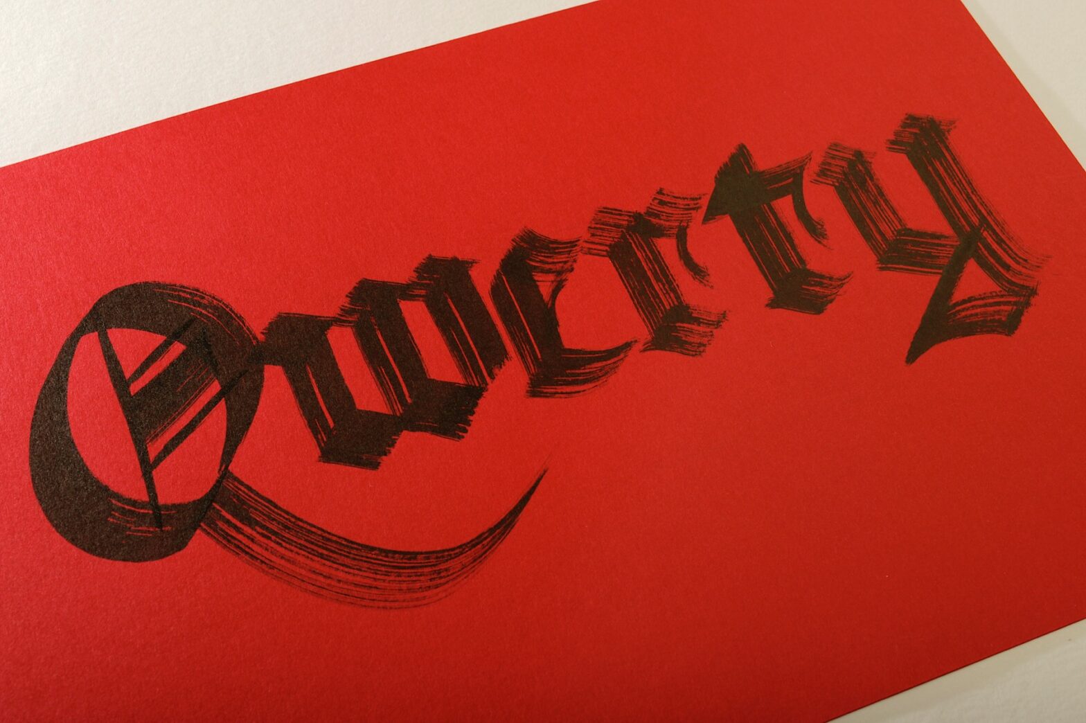 Trabalho de caligrafia em preto sobre papel vermelho, onde está escrito “Qwerty” em letras góticas