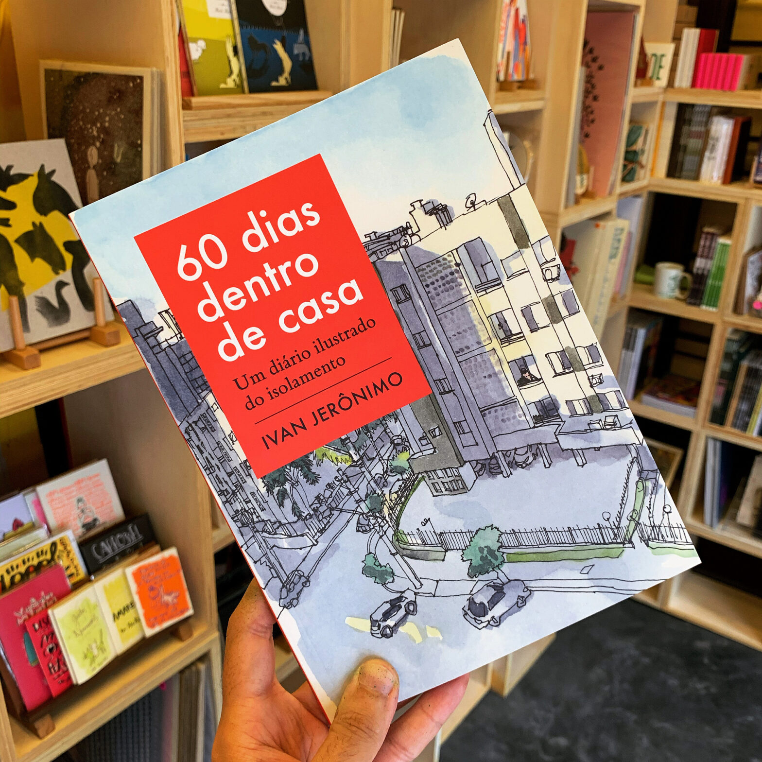 Foto de mão segurando o livro “60 dias dentro de casa – Um diário ilustrado do isolamento” entre as estantes no interior da Banca Curva
