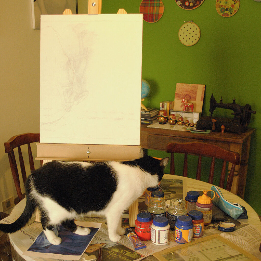 cavalete de mesa com tela já esboçada a lápis. Gata preto e branca caminha entre os potes de tinta