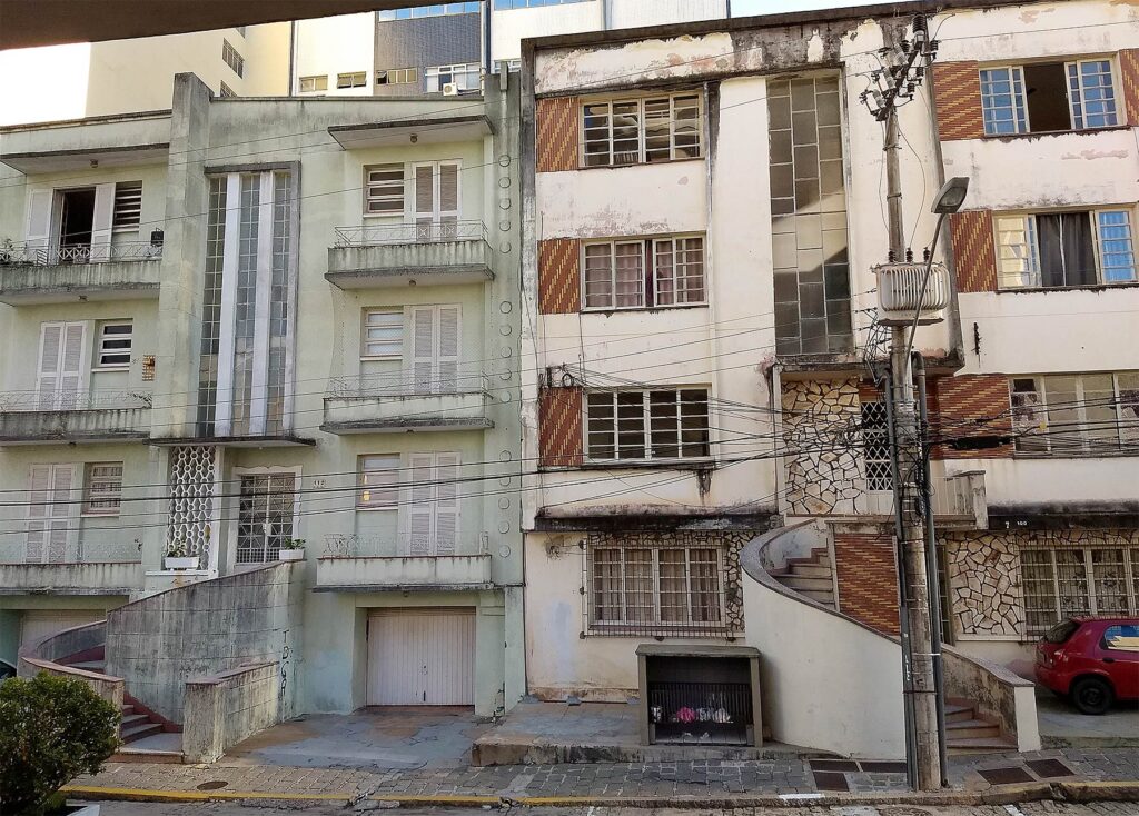 Foto dos dois prédios. O da esquerda é verde claro, enquanto o da direita é creme com detalhes em cerâmica vermelha. Ambos têm quatro pavimentos