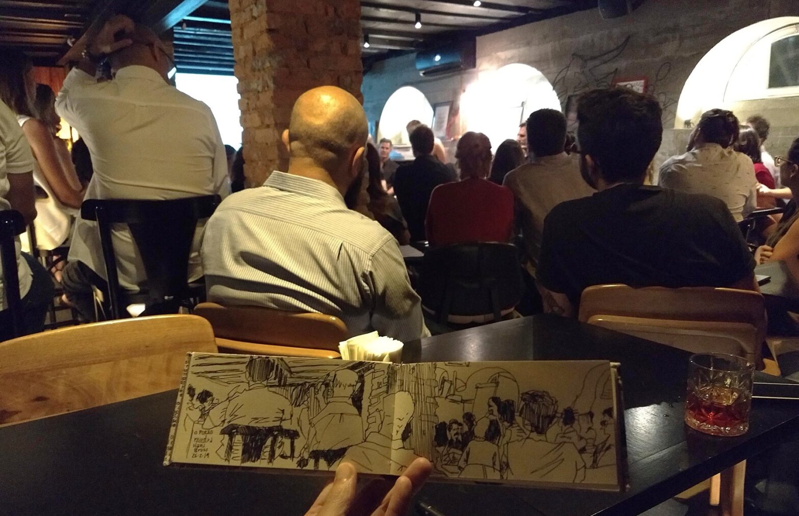 Público do evento no Porão, virado para os palestrantes. Em primeiro plano, caderno horizontal aberto com o desenho