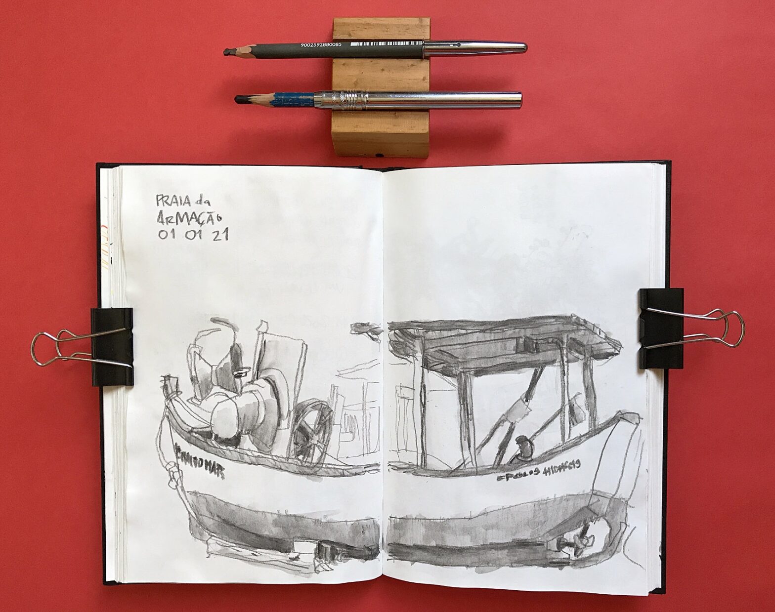 Foto de caderno aberto com desenho em página dupla de um barco de pesca. Acima do caderno, dois lápis.