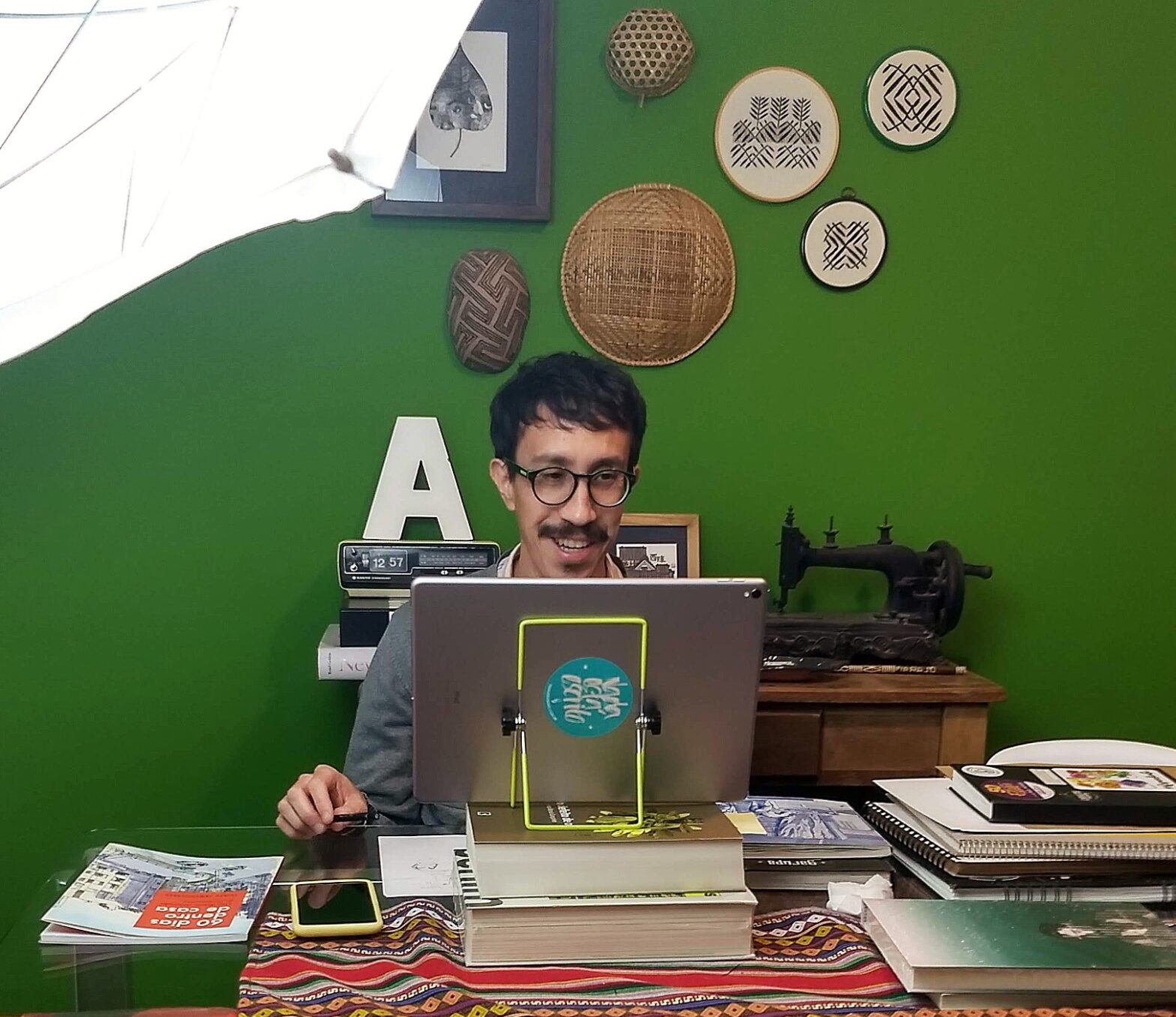 Foto do autor sentado em uma mesa, em frente a um tablet, cercado por cadernos, com uma parede verde ao fundo, onde estão pendurados bordados e objetos indígenas