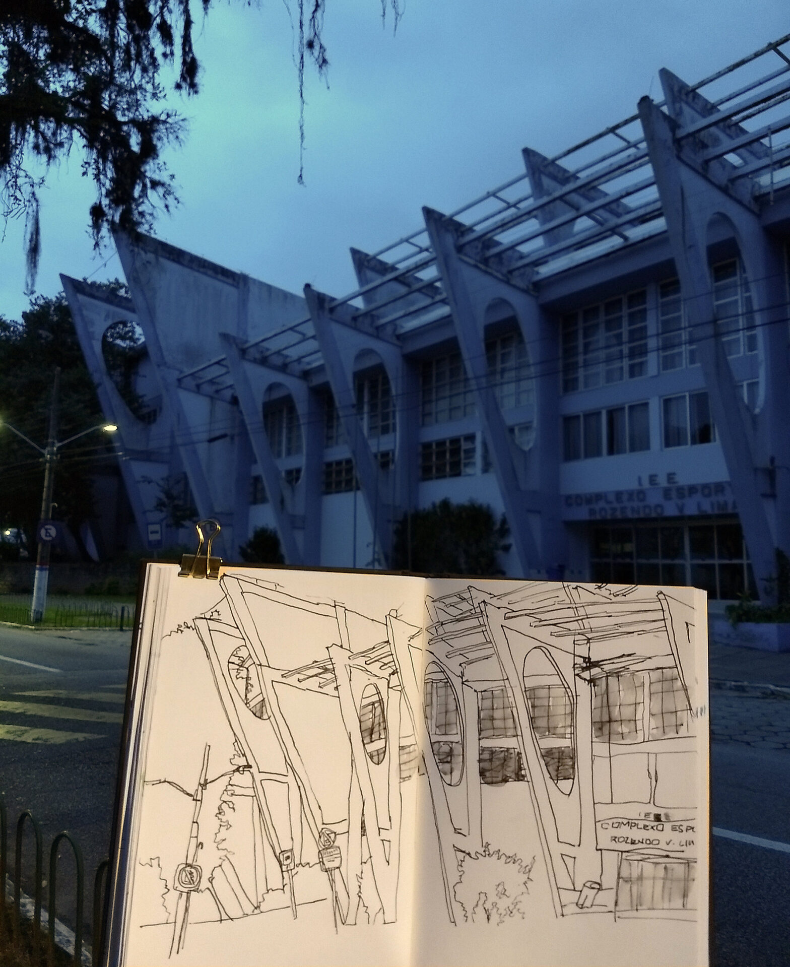 Em primeiro plano, caderno com desenho da fachada do ginásio. Ao fundo, o ginásio verdadeiro, com as colunas triangulares