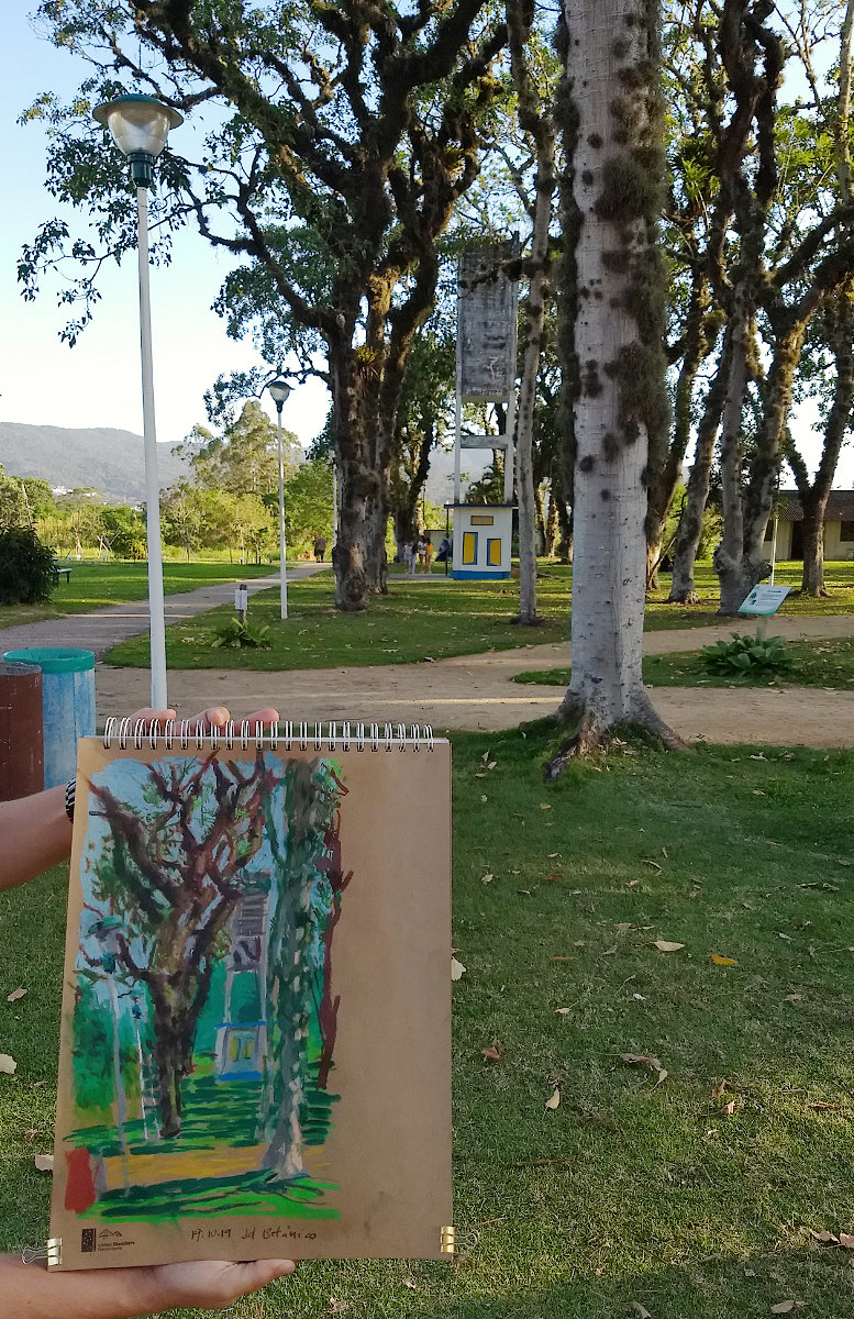 Cena do Jardim Botânico de Florianópolis em um dia ensolarado com árvores e uma caixa d'água ao fundo. Em primeiro plano, mãos seguram caderno com pintura me pastel seco