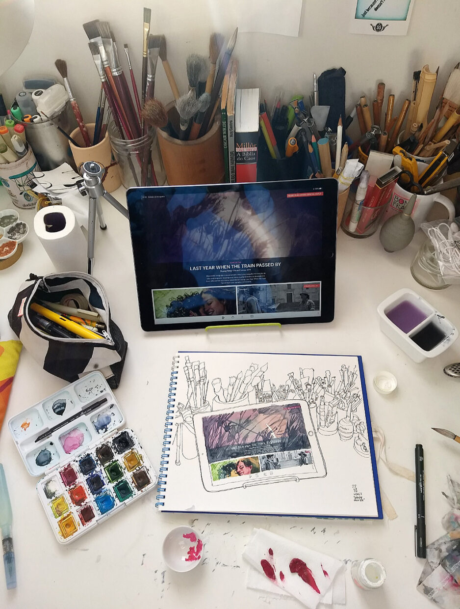 Mesa de desenho com o caderno, um tablet inclinado e vários materiais de desenho e pintura