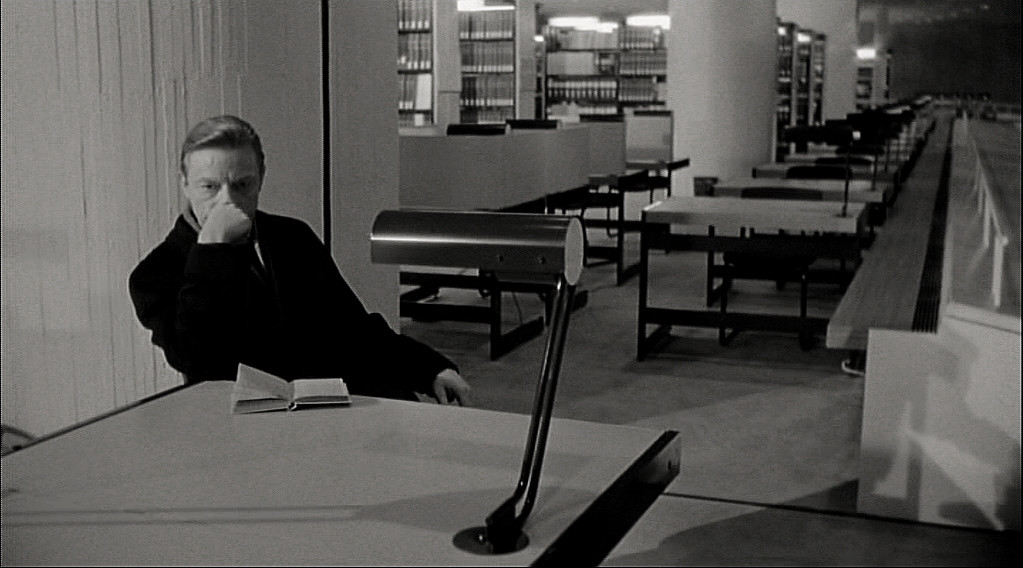 Cena do filme em que o personagem está na sentado em uma mesa na biblioteca com outras mesas e prateleiras ao fundo