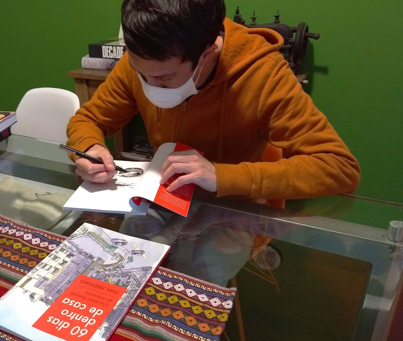 Autor de casaco ocre em uma mesa de vidro escrevendo a dedicatória em um livro aberto. À sua frente, outro livro fechado. Ao fundo, parede verde