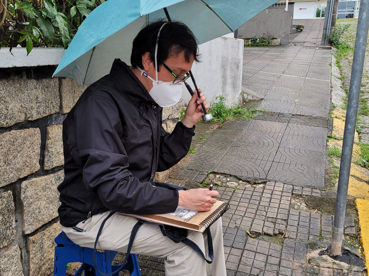 Autor de casaco preto desenhando sentado em uma banqueta na calçada com o caderno no colo e um guarda-chuva azul na mão esquerda