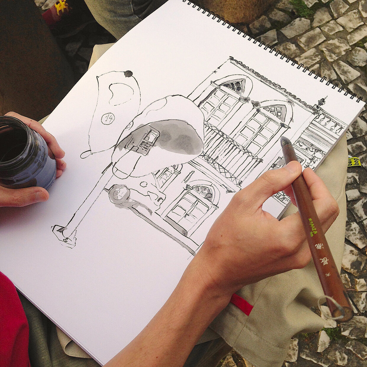 Fotografia do autor com o caderno apoiado no colo, com o desenho quase pronto, segurando o pincel e um pote de tinta