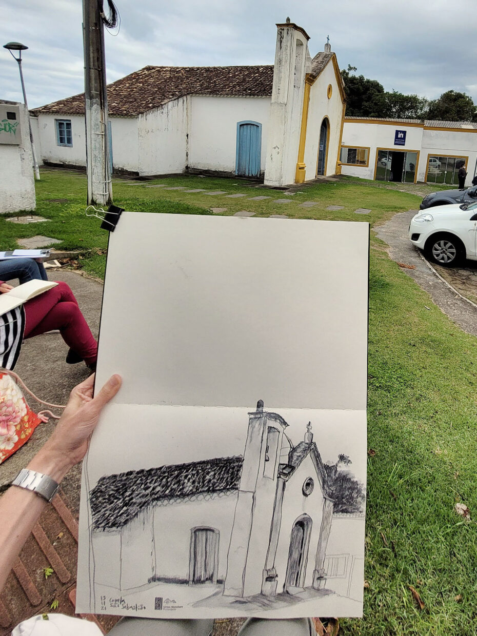 Foto com a capela ao fundo e em primeiro plano caderno aberto com o desenho