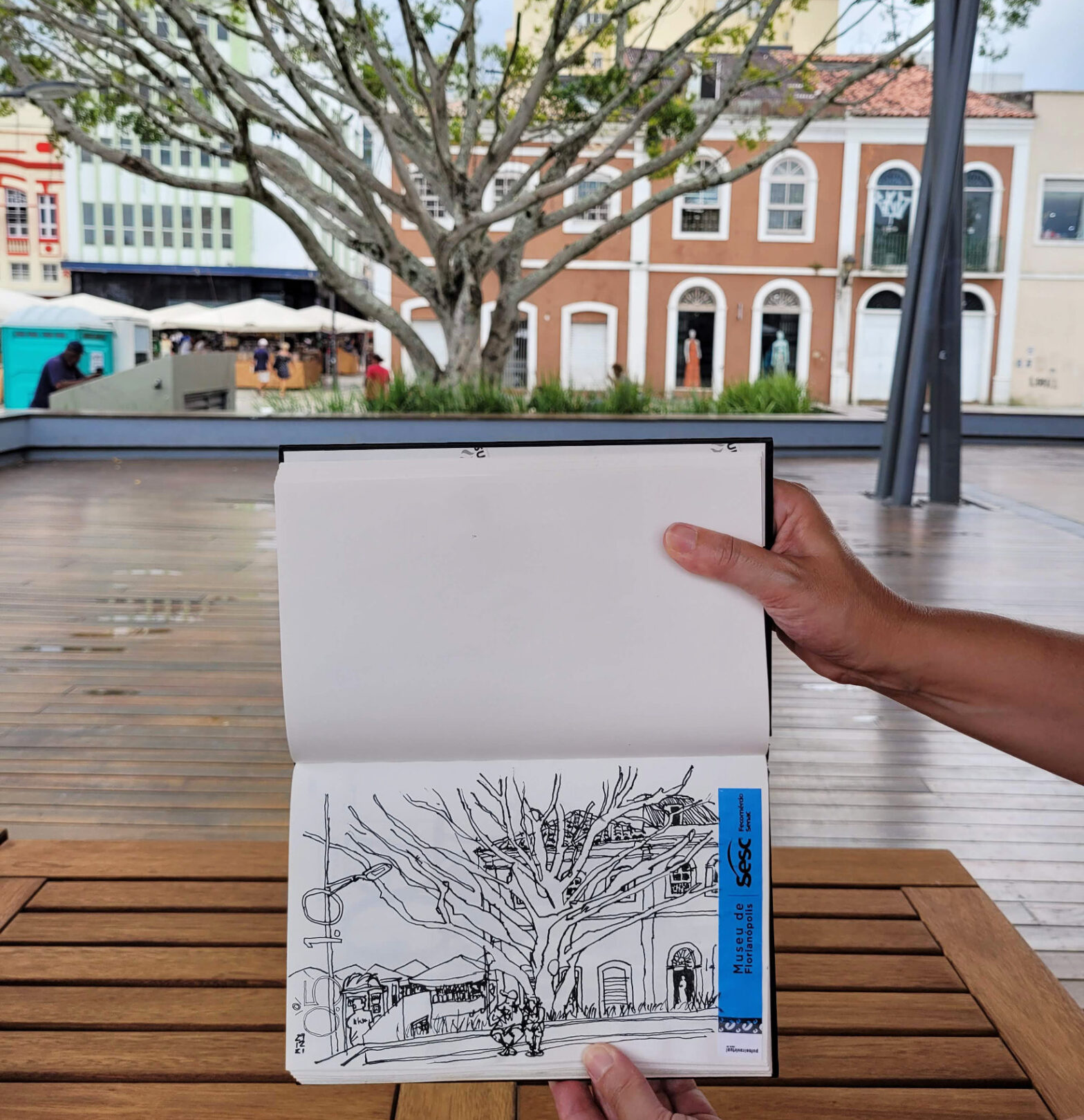 Foto mostrando o caderno aberto com o desenho. Ao fundo, o cenário retratado: mureta, árvore e casarões do comércio do Largo da Alfândega