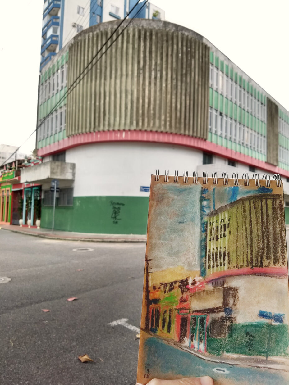 Foto com o prédio da Diretoria de Saúde do Servidor de Santa Catarina ao fundo com o caderno com o desenho em primeiro plano