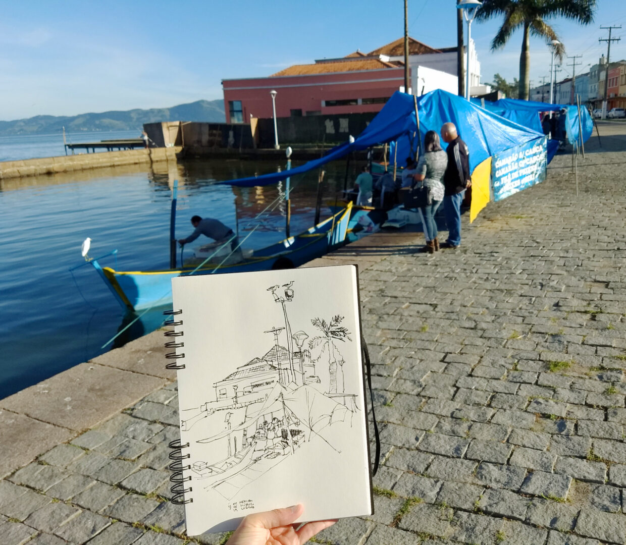 Foto do caderno em primeiro plano com barco e tenda de do peixeiro amarrada na doca