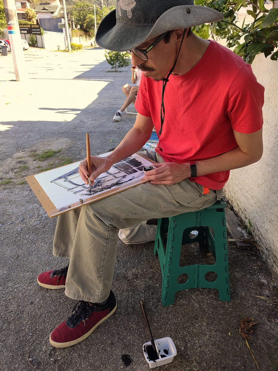 Autor sentado em um banco desenha em cima de uma prancheta apoiada em seu colo
