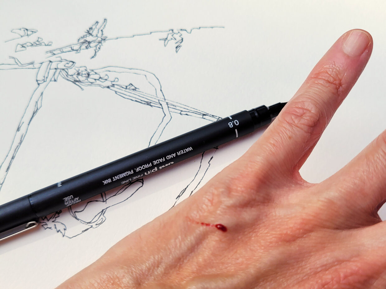 Parte do papel com desenho, uma caneta e a mão do autor com picada de mosquito saindo sangue
