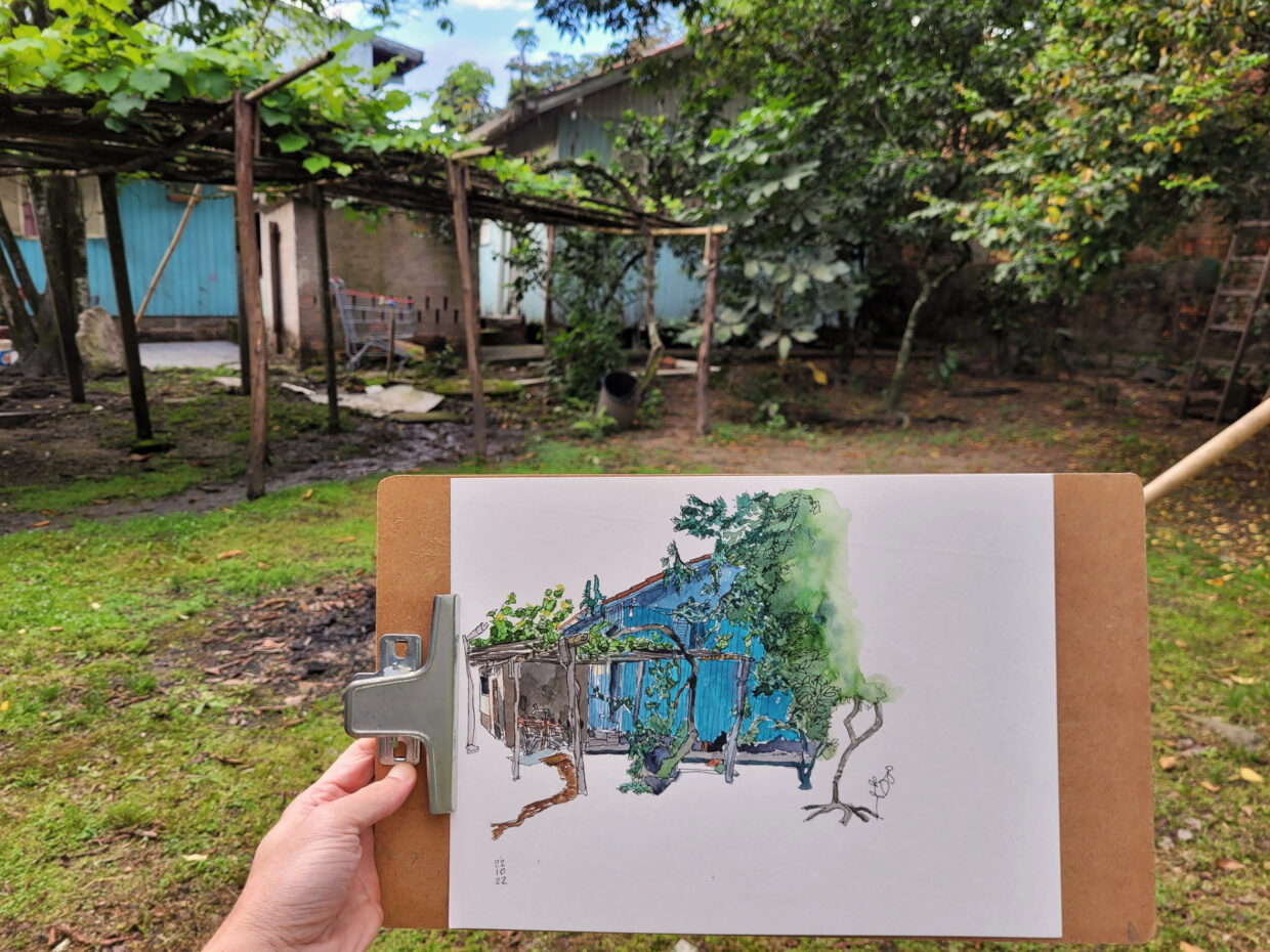 Terreno com parreira e duas casas de madeira. Autor segura prancheta e desenho em primeiro plano