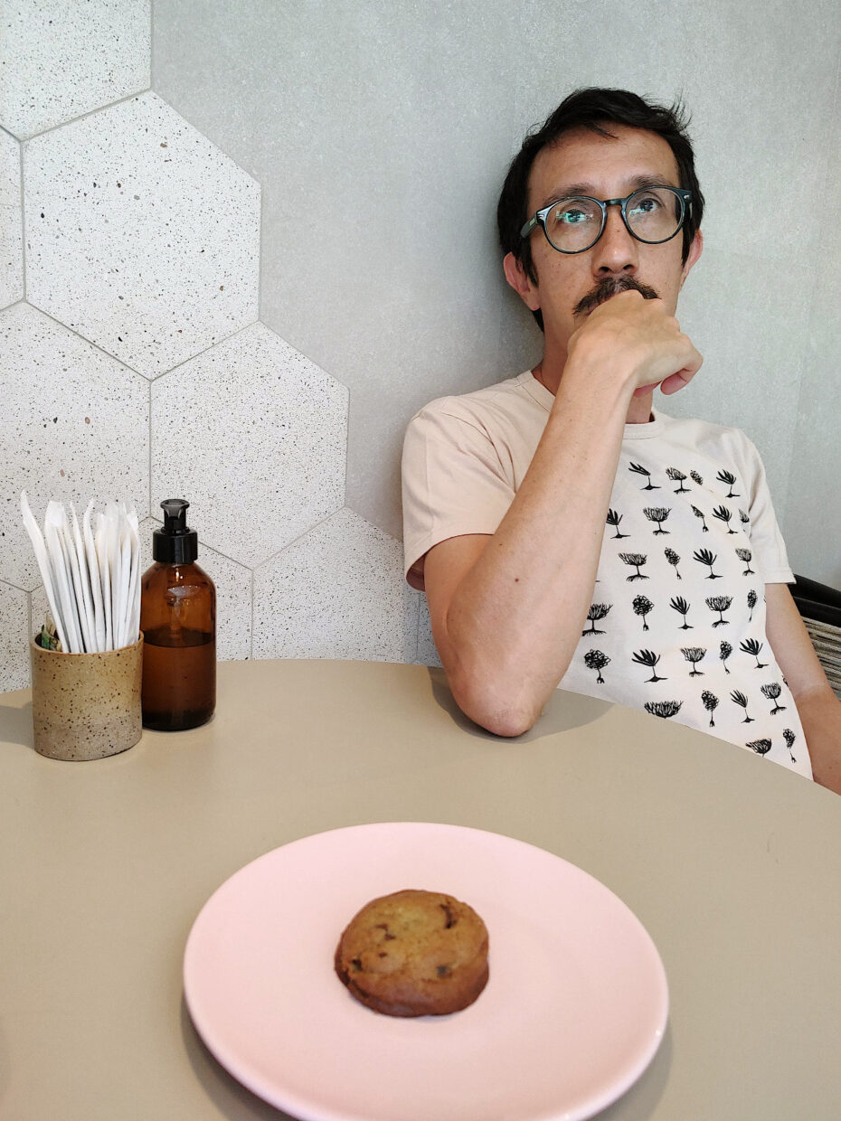 Autor Ivan Jerônimo usando óculos e camiseta com estampa de sementes, sentado em uma mesa com um cookie à sua frente