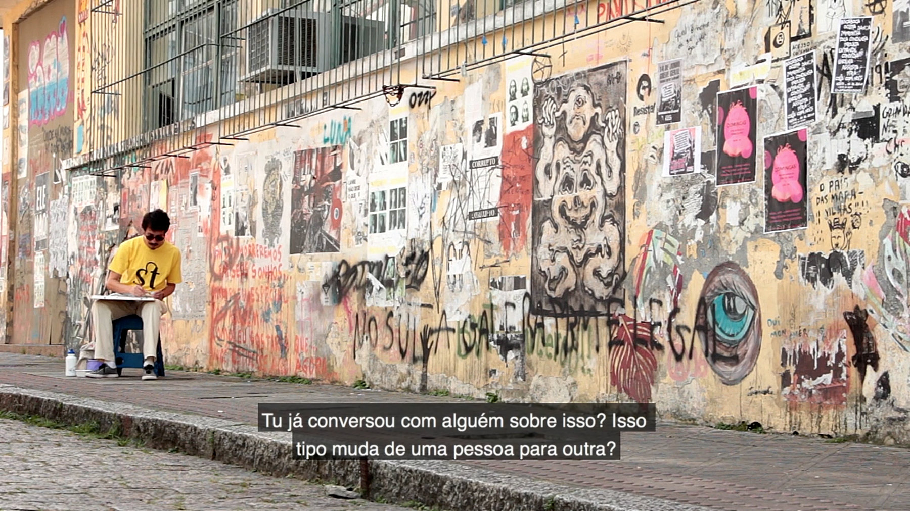 Quadro do documentário com Ivan Jerônimo sentado, desenhando ao lado da fachada do a antiga escola Antonieta de Barros