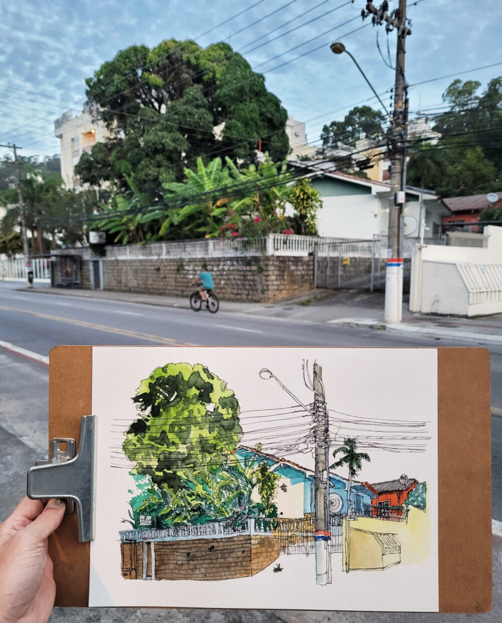Foto da prancheta com o desenho em primeiro plano. Ao fundo, a rua do Córrego Grande com as casas retratadas e um homem passando de bicicleta