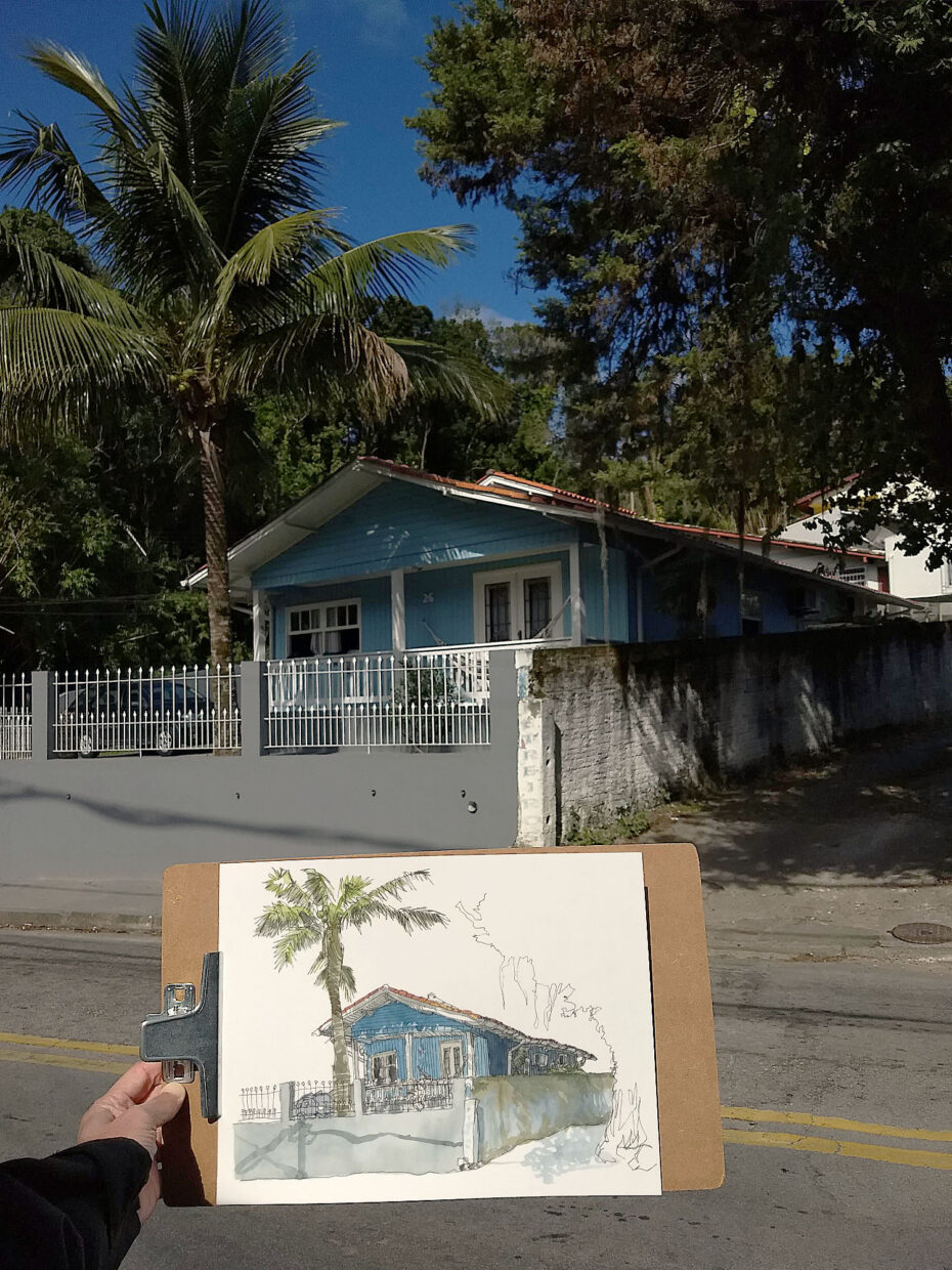 Fotografia da mão do autor segurando o desenho da casa de madeira azul em primeiro plano, com a casa de fundo