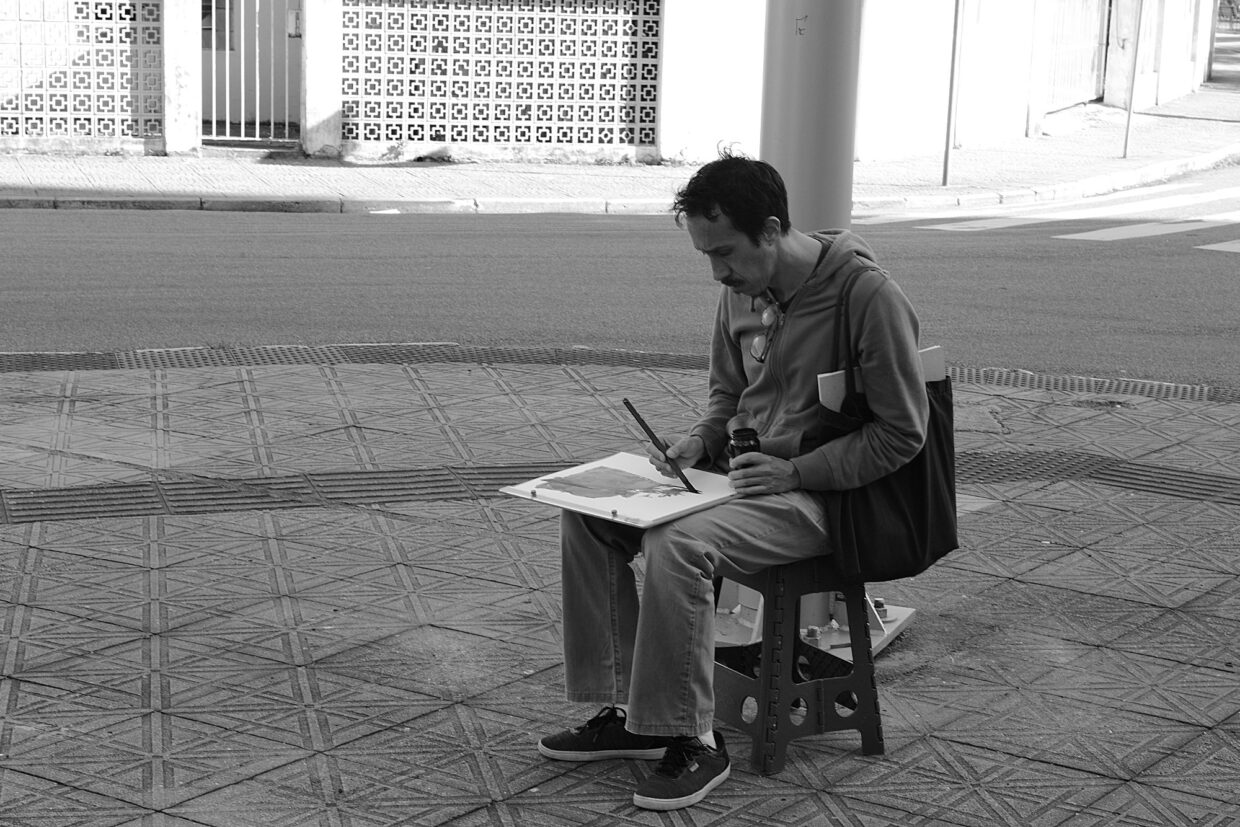 Foto em preto e branco mostrando o autor, Ivan Jerônimo, sentado na calçada, desenhando com uma prancheta no colo