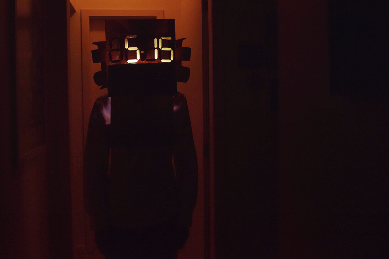 Fotografia de uma pessoa sob o batente de uma porta com uma máscara de papel onde está indicada a hora “5:15”