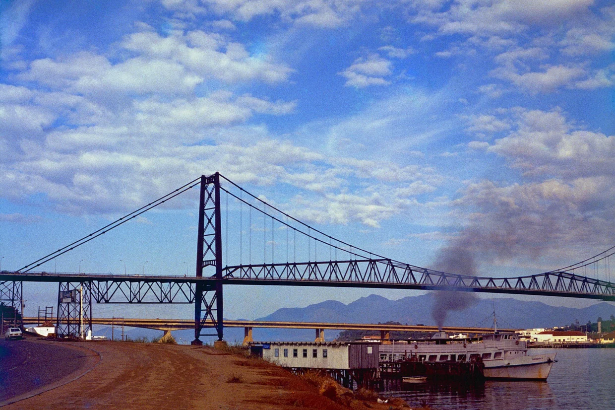 Foto da ponte Hercílio Luz na década de 1980, ainda pintada de preto, com a ponte Colombo Sales ao fundo. Em primeiro plano, a avenida Beira-Mar sem calçada nem ciclovia e um barco ancorado chamado Turimar V