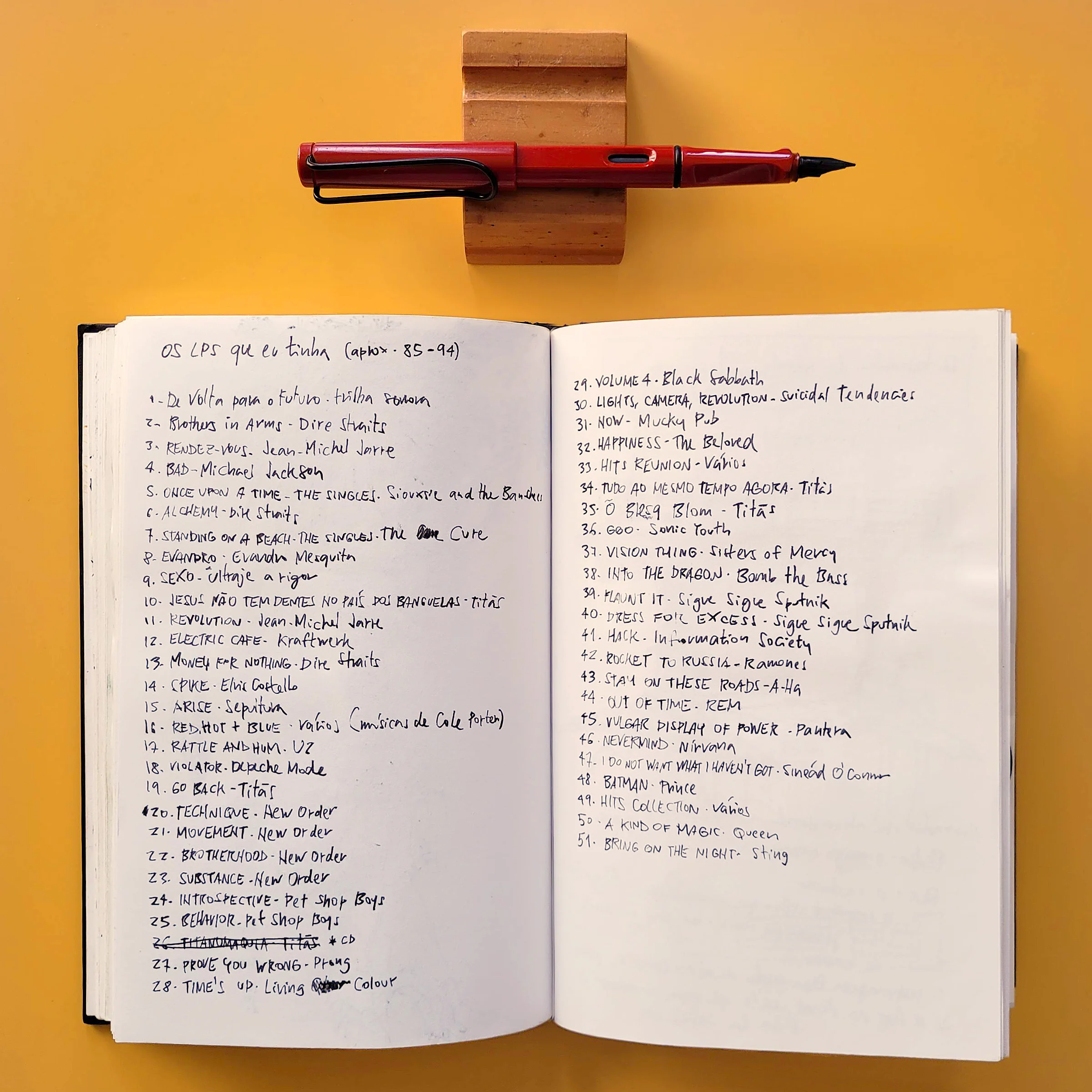Foto de um caderno aberto com uma lista de discos (álbuns, LPs) sobre uma mesa laranja. Acima, uma caneta descansa sobre um suporte¸