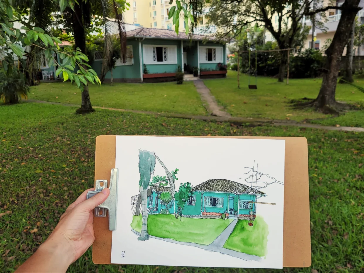 Fotografia do quintal com a casa tendo em primeiro plano a mão do autor segurando o desenho da mesma casa