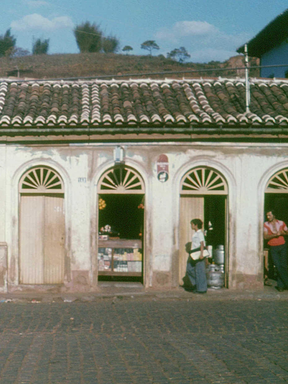 Fotografia antiga de um menino andando em frente a uma mercearia numa construção mais antiga ainda