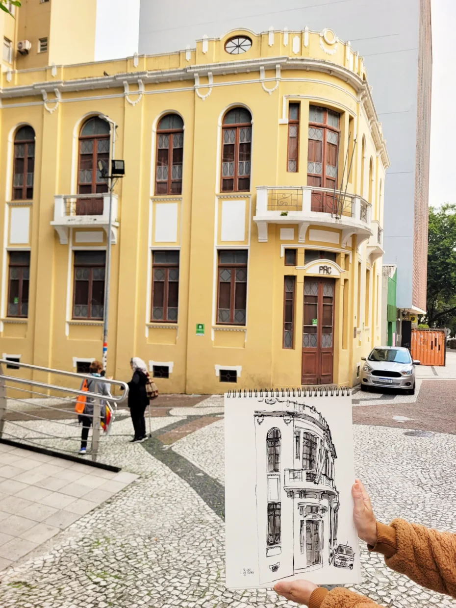 Fotografia da Casa da Memória, um prédio de arquitetura eclética, de esquina, pintado de amarelo. Em primeiro plano, um caderno com o desenho do prédio