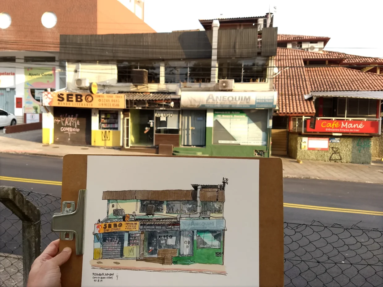 Fotografia mostrando em primeiro plano uma prancheta com o desenho do imóvel com vários estabelecimentos (sebo, café e birô gráfico). Ao fundo, a cena real na luz de fim de tarde