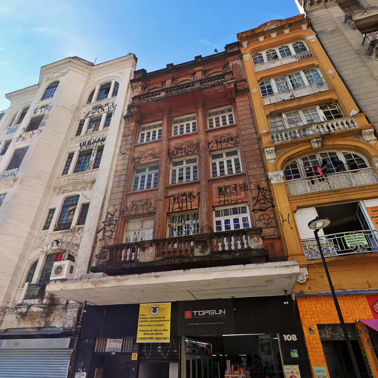 Fachada pichada e um tanto envelhecida, de tom vermelho, do prédio onde fica o Estúdio Lâmina. O quarto andar, que abriga o estúdio, tem sacada e janelas arredondadas.