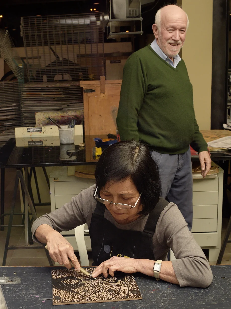 Julia Iguti sentada em uma mesa gravando o desenho em uma matriz de madeira, tendo ao fundo Antônio C. Silva em pé. Estão no ateliê de gravura do CIC, onde se veem vários equipamentos