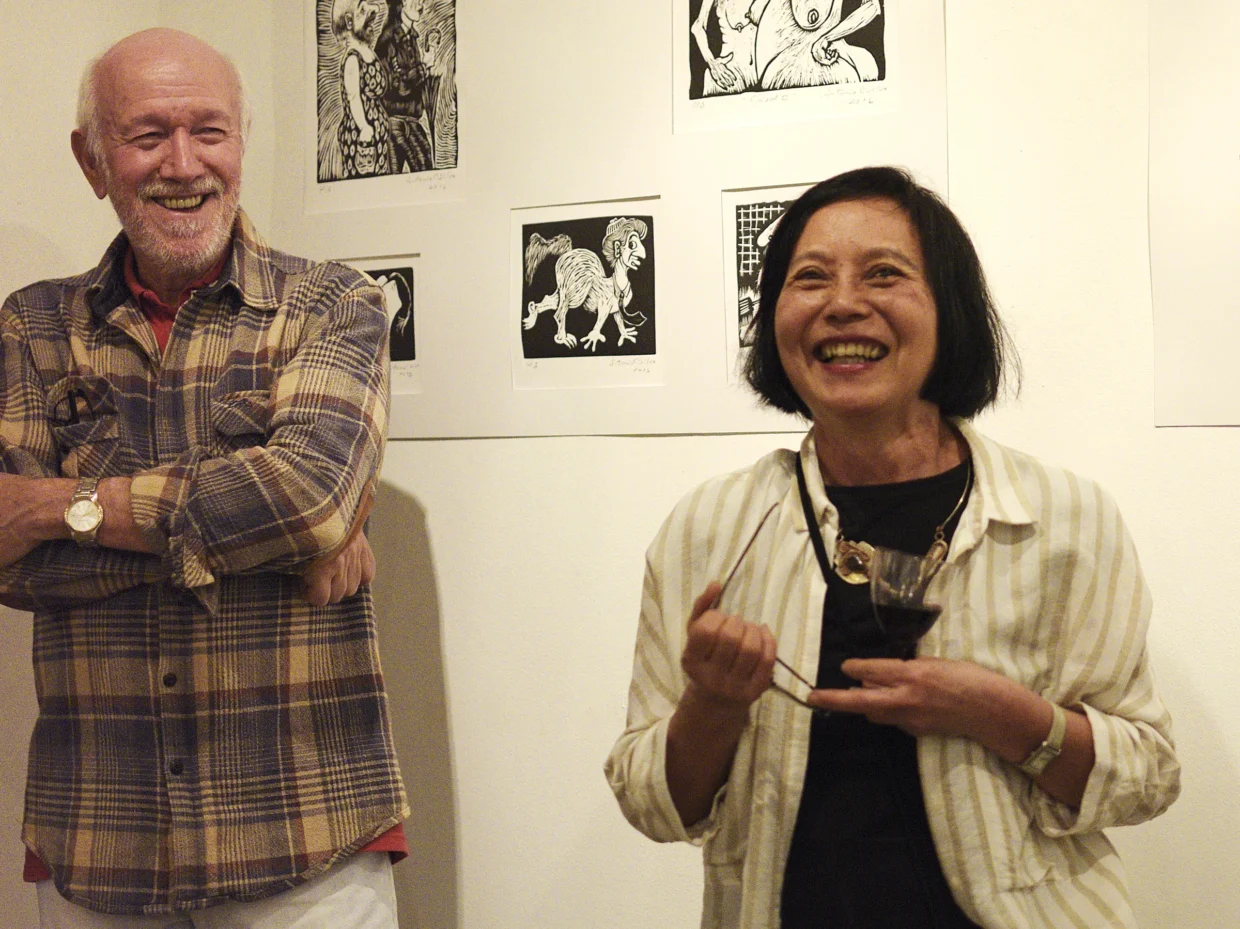 Artistas Antonio C. Silva (à esquerda) e Julia Iguti (direita) em pé, sorridentes, tendo ao fundo a parede de uma exposição com xilogravuras