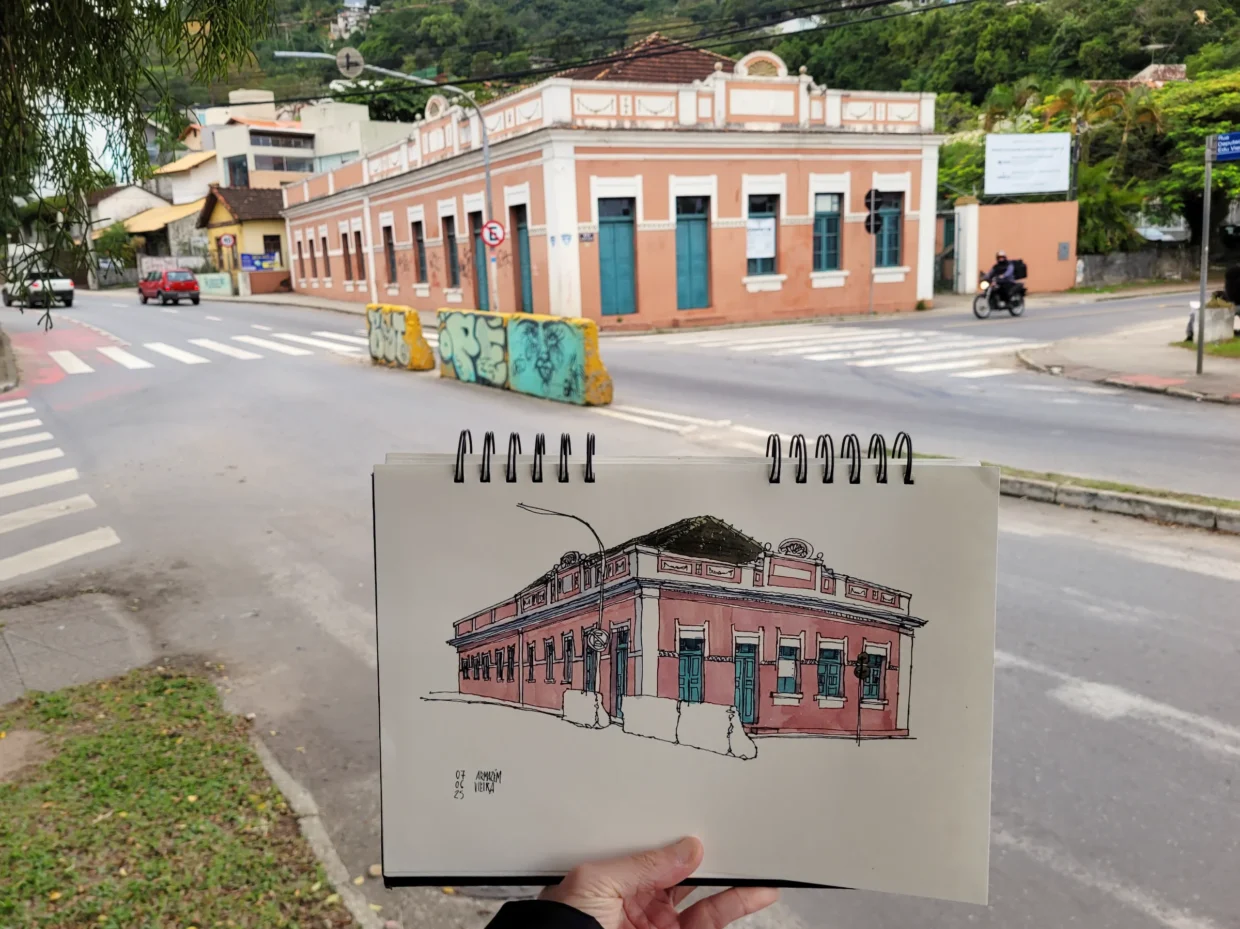 Fotografia mostrando o autor segurando o caderno com o desenho do Armazém Vieira em primeiro plano. Ao fundo, o próprio Armazém e os blocos de concreto, grafitados, no cruzamento das ruas
