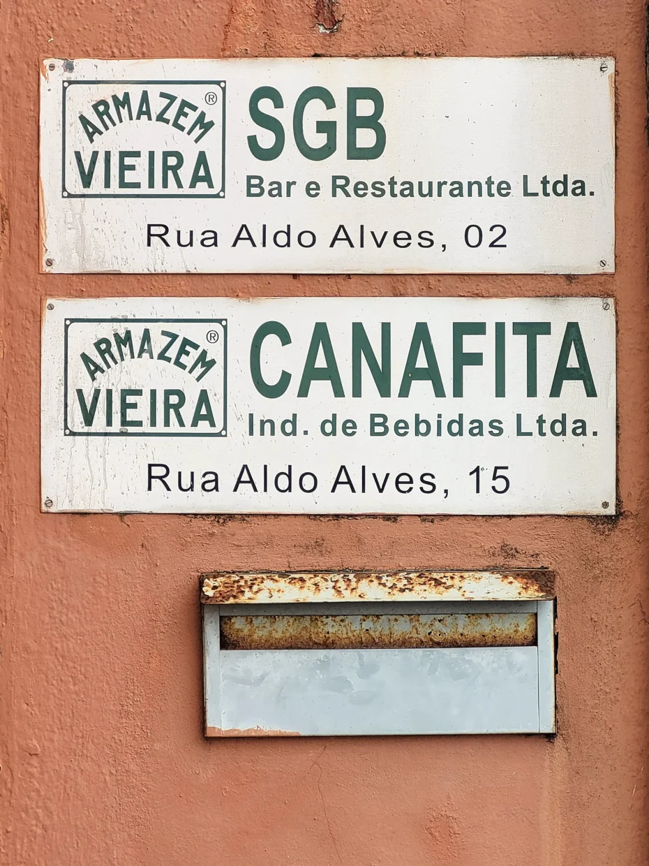 Foto mostrando caixa de correio e duas placas do Armazém Vieira fixadas próximas à entrada: - SGB - Bar e Restaurante Ltda. Rua Aldo Alves, 02 - CANAFITA - Ind. de Bebidas Ltda. Rua Aldo Alves, 15