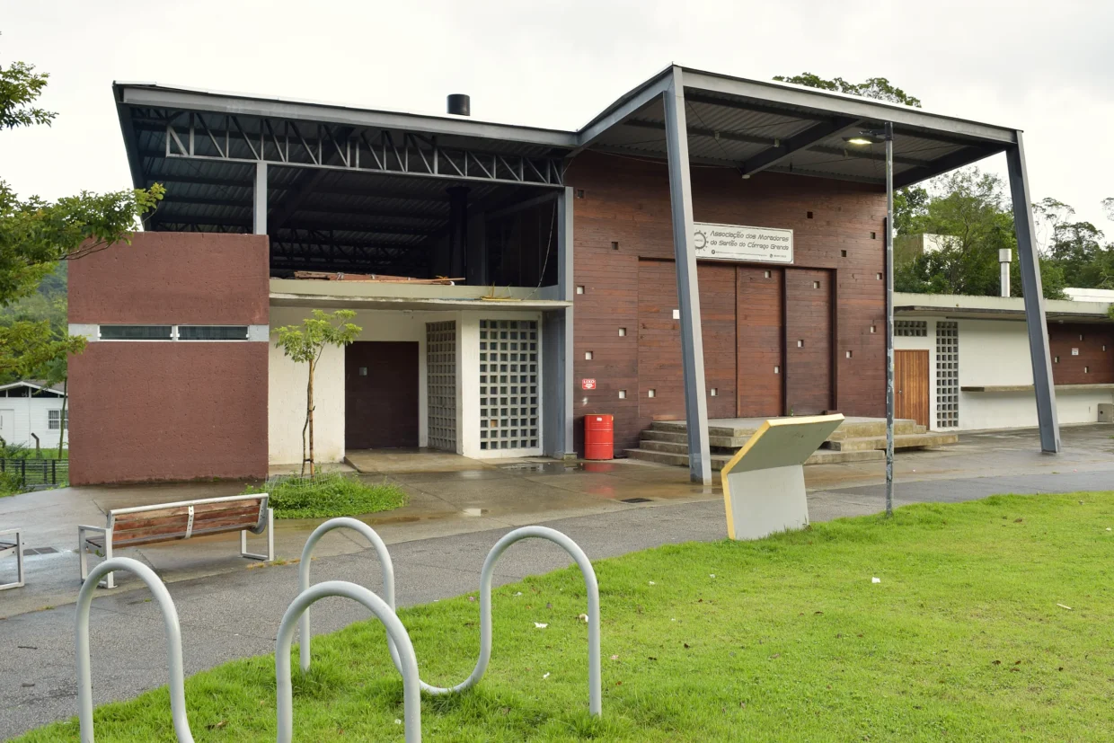 Fachada do pavilhão comunitário da Associação dos Moradores do Sertão do Córrego Grande, com paineis e madeira e estrutura metálica aparente. Em frente, gramado da praça
