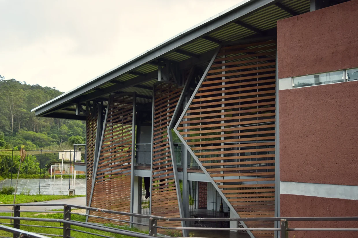 Fundos do pavilhão comunitário com elementos metálicos com grades de madeira. Ao fundo, uma quadra polivalente de cimento