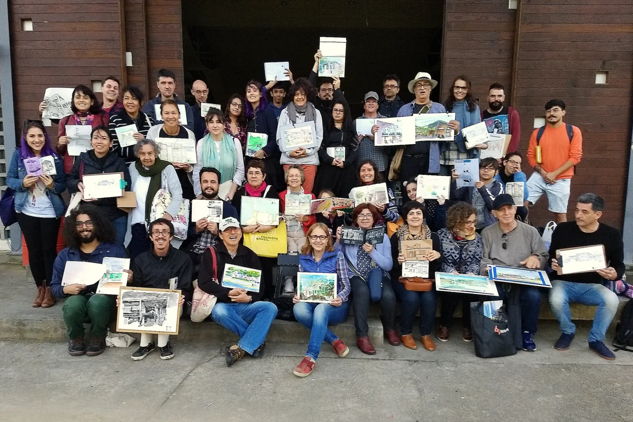 Participantes do encontro do URban Sketchers Florianópolis agrupados para a foto, segurando seus desenhos. Estão em frente ao portão principal do pavilhão da Amosc