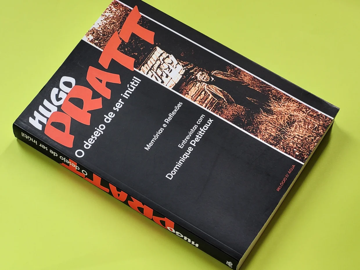 Foto de capa do livro “Hugo Pratt - O desejo de ser inútil“, onde está escrito: Memórias e Reflexões - Entrevistas com Dominique Petitfaux - ed. Relógio D'Água. A capa é preta com letras vermelhas e brancas, com uma foto vertical de Pratt, já na meia idade, num campo, com um casebre ao fundo.