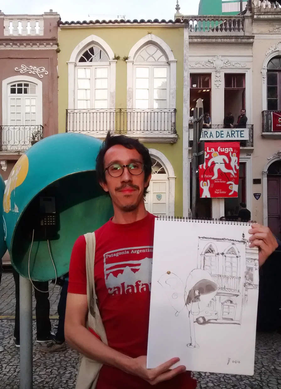 Autor Ivan Jerônimo, de camiseta vermelha, segura o caderno com o desenho mostrando o casario antigo e um orelhão (telefone público) em primeiro plano