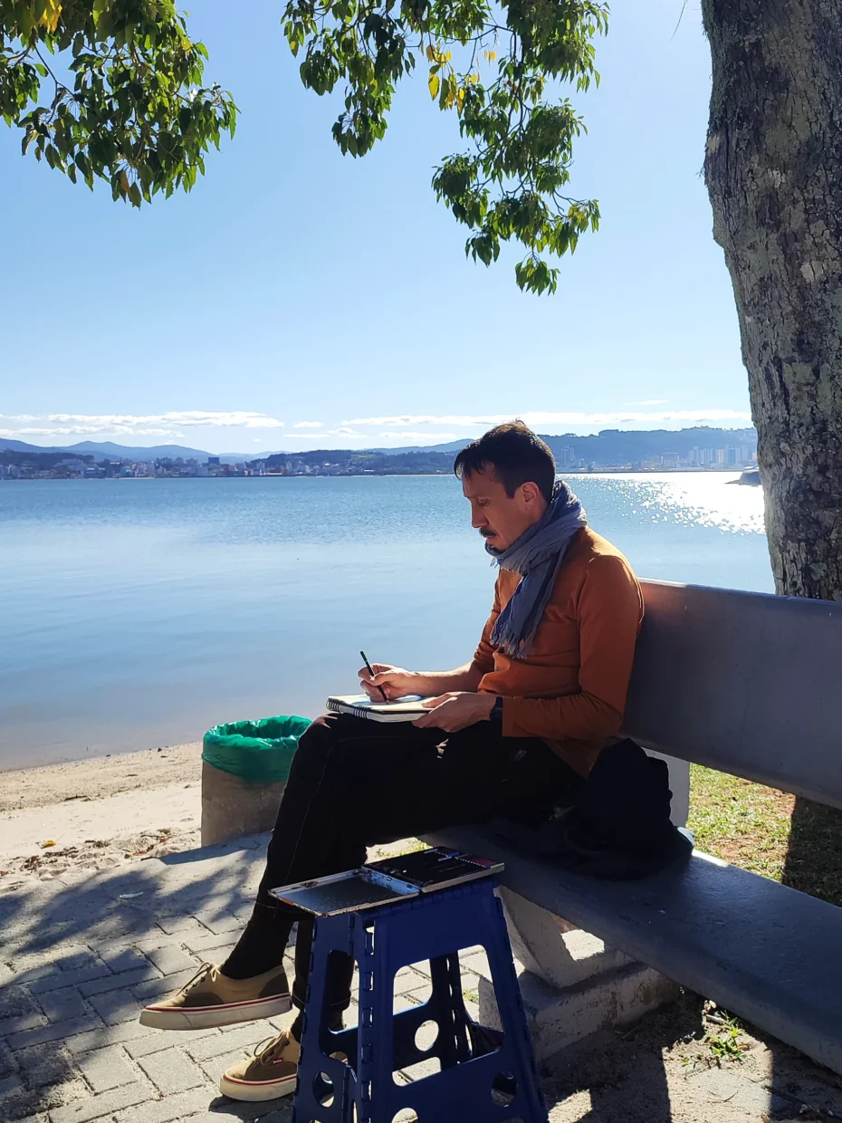 O autor, Ivan Jerônimo, sentado em um banco à beira do mar, desenhando com o caderno no colo